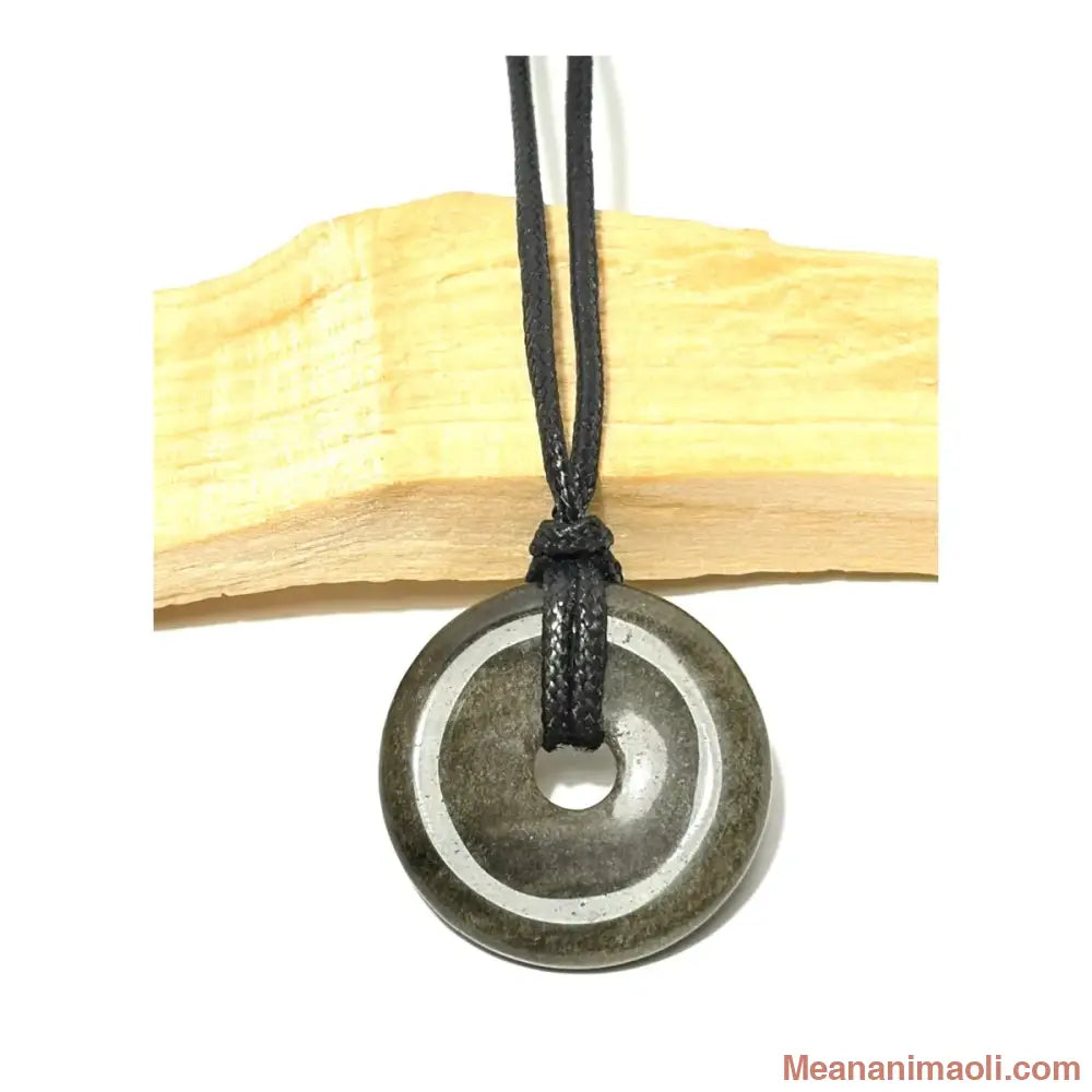 Pendentif donut en Obsidienne Dorée naturelle aux reflets dorés suspendu par un cordon en coton ciré noir devant un morceau de bois de Palo Santo - MEANANIMAOLI
