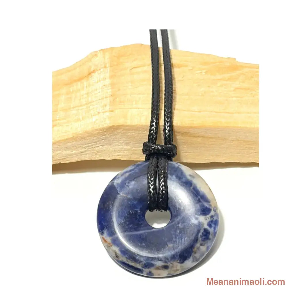 Pendentif donut en Sodalite naturelle bleu nuit et blanc suspendu par un cordon en coton ciré noir devant un morceau de bois de Palo Santo - MEANANIMAOLI