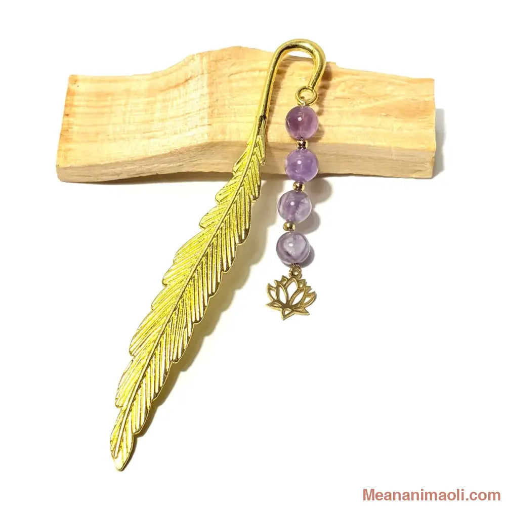 Marque-page en forme de plume dorée avec quatre perles rondes en améthyste violette et une breloque en forme de fleur de lotus dorée, posé sur un bâton de bois clair.