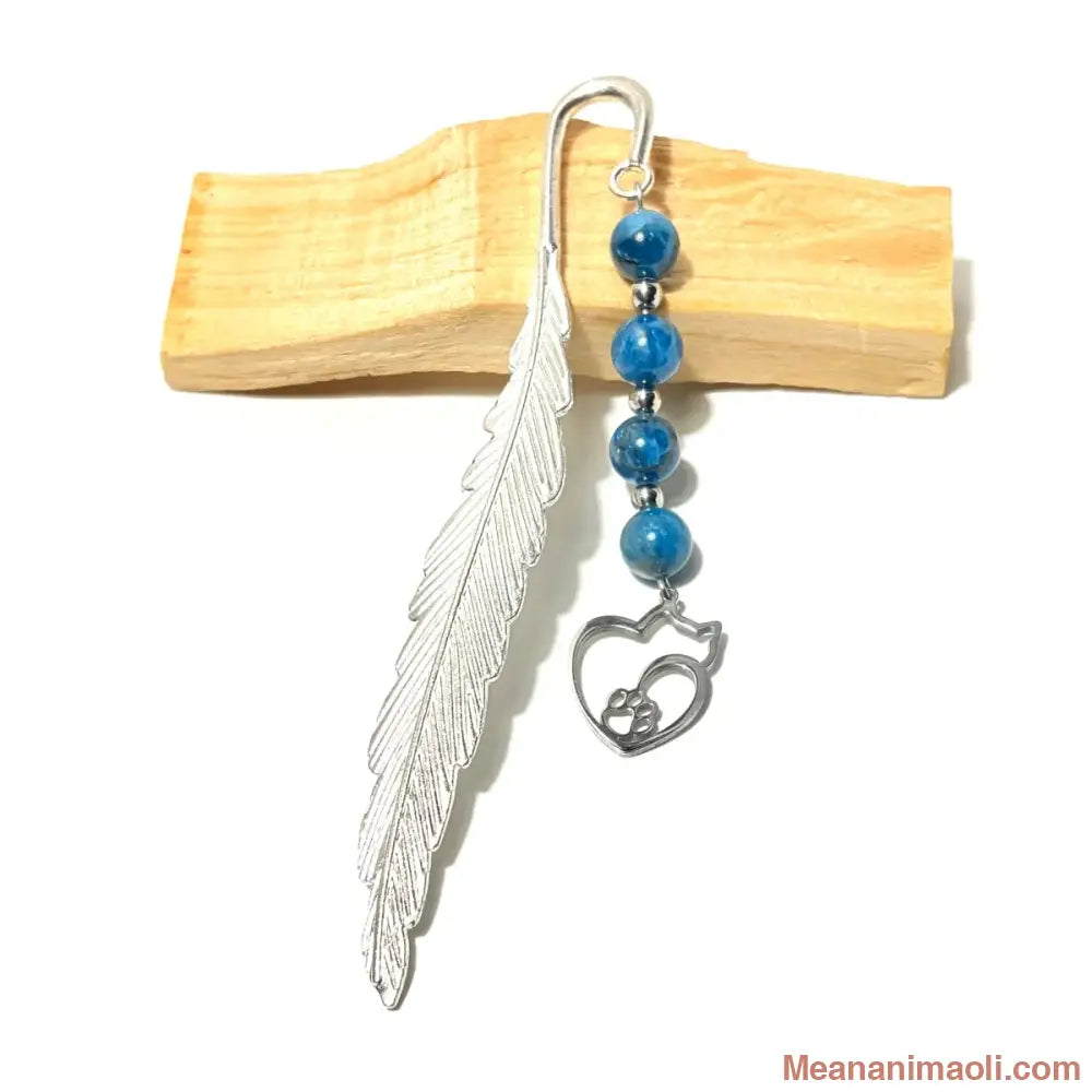 Marque-page en forme de plume argentée avec quatre perles rondes en apatite bleue et une breloque en forme  de chat stylisée contenant une petite patte, présenté sur un bâton de bois clair.