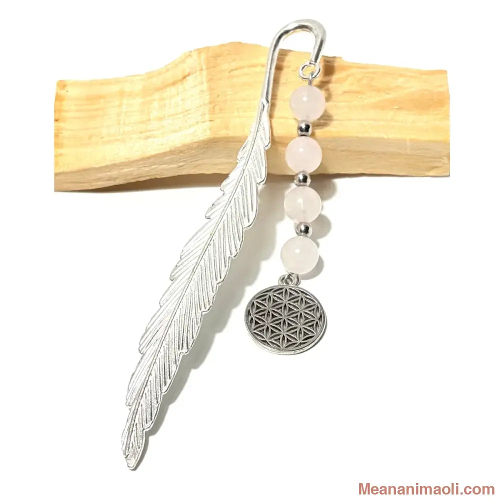 Marque-page en forme de plume argentée orné de quatre perles rondes en quartz rose et d'une breloque ronde représentant le symbole de la Fleur de Vie, posé sur un morceau de bois de Palo Santo.