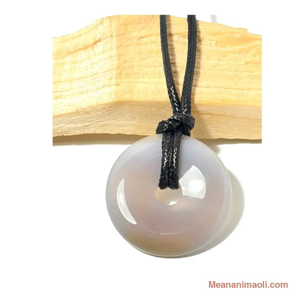 Pendentif donut en Agate naturelle gris clair suspendu par un cordon en coton ciré noir devant un morceau de bois de Palo Santo - MEANANIMAOLI