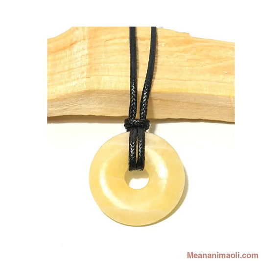 Pendentif donut en Calcite Jaune naturelle couleur miel suspendu par un cordon en coton ciré noir devant un morceau de bois de Palo Santo - MEANANIMAOLI